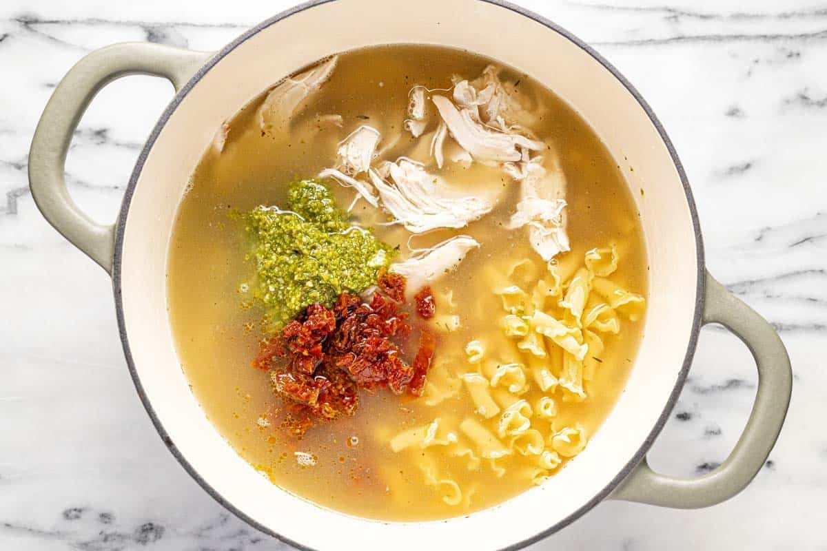 Large pot with stock, chicken, pesto, sun-dried tomatoes, and noodles. 