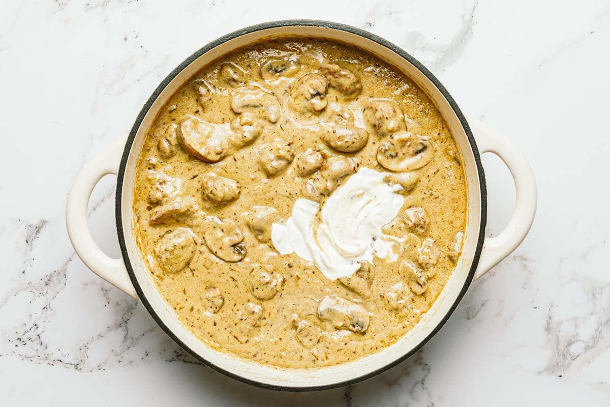 Sour cream being added to a large pan of meatball stroganoff. 