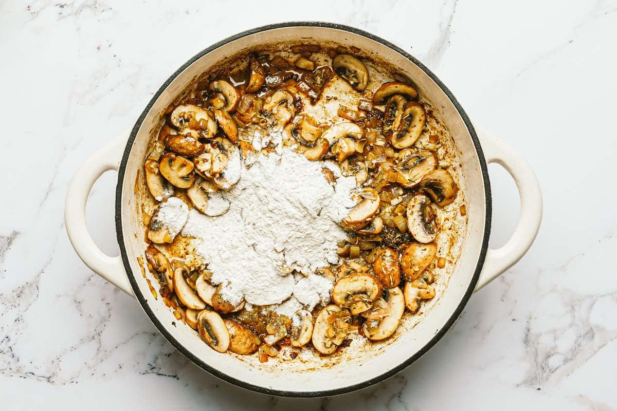 Sliced mushrooms, onions, herbs, and flour in a large pan. 