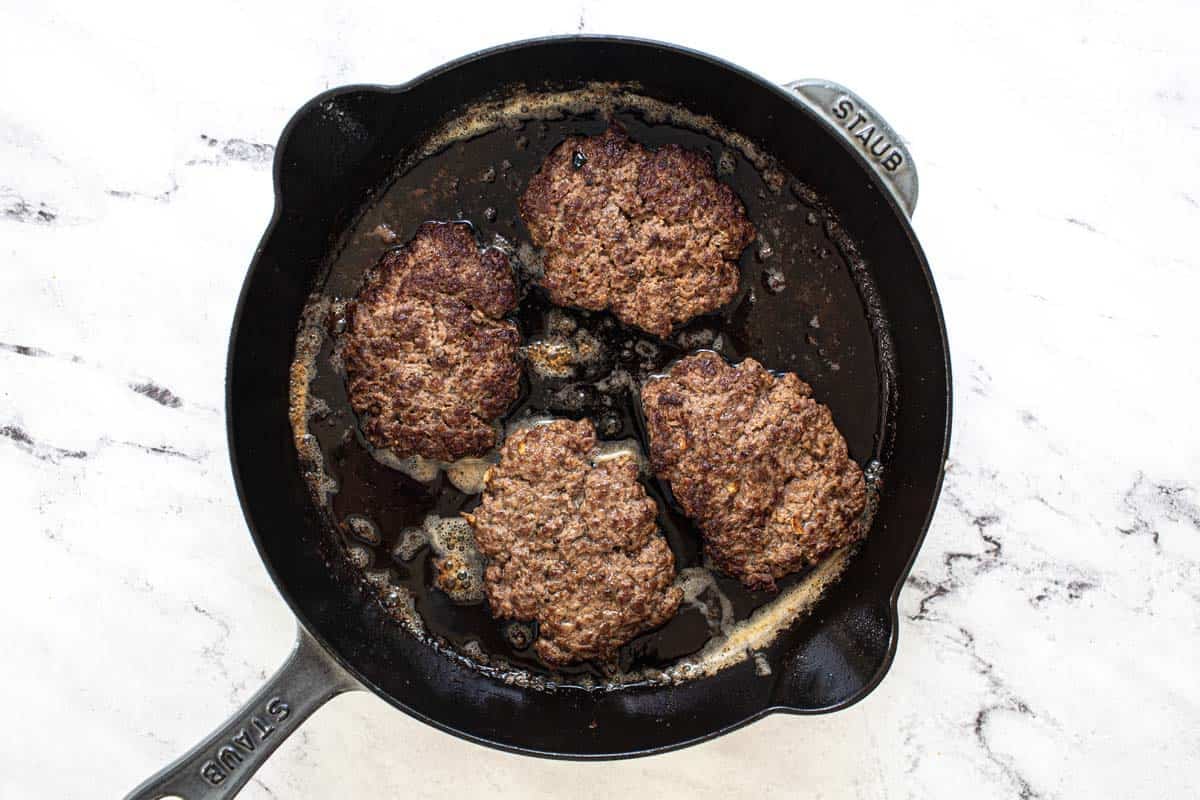 Cast iron pan with cooked ground beef patties.