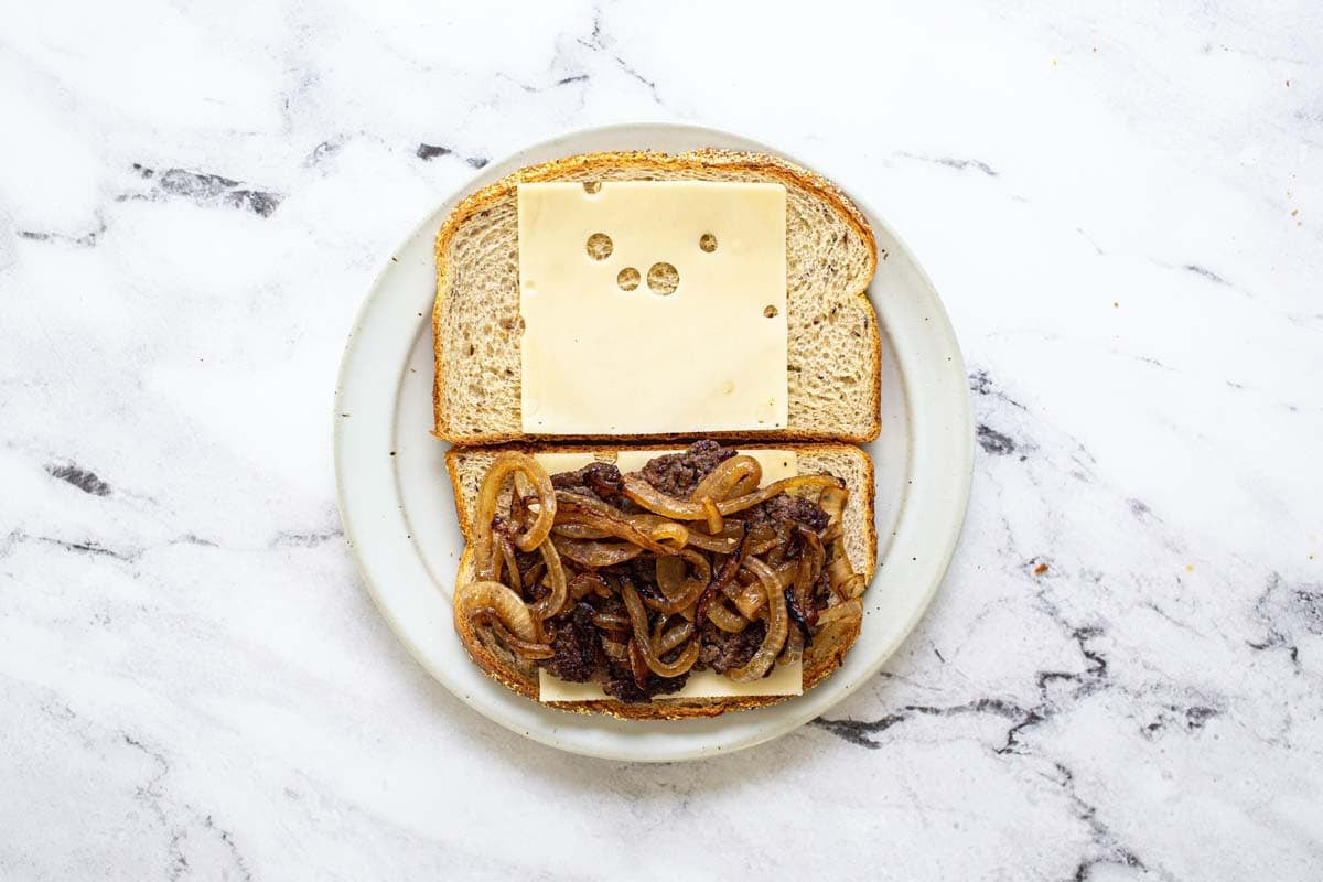 Two pieces of rye bread on a plate with sliced cheese, burger patty, and caramelized onions.