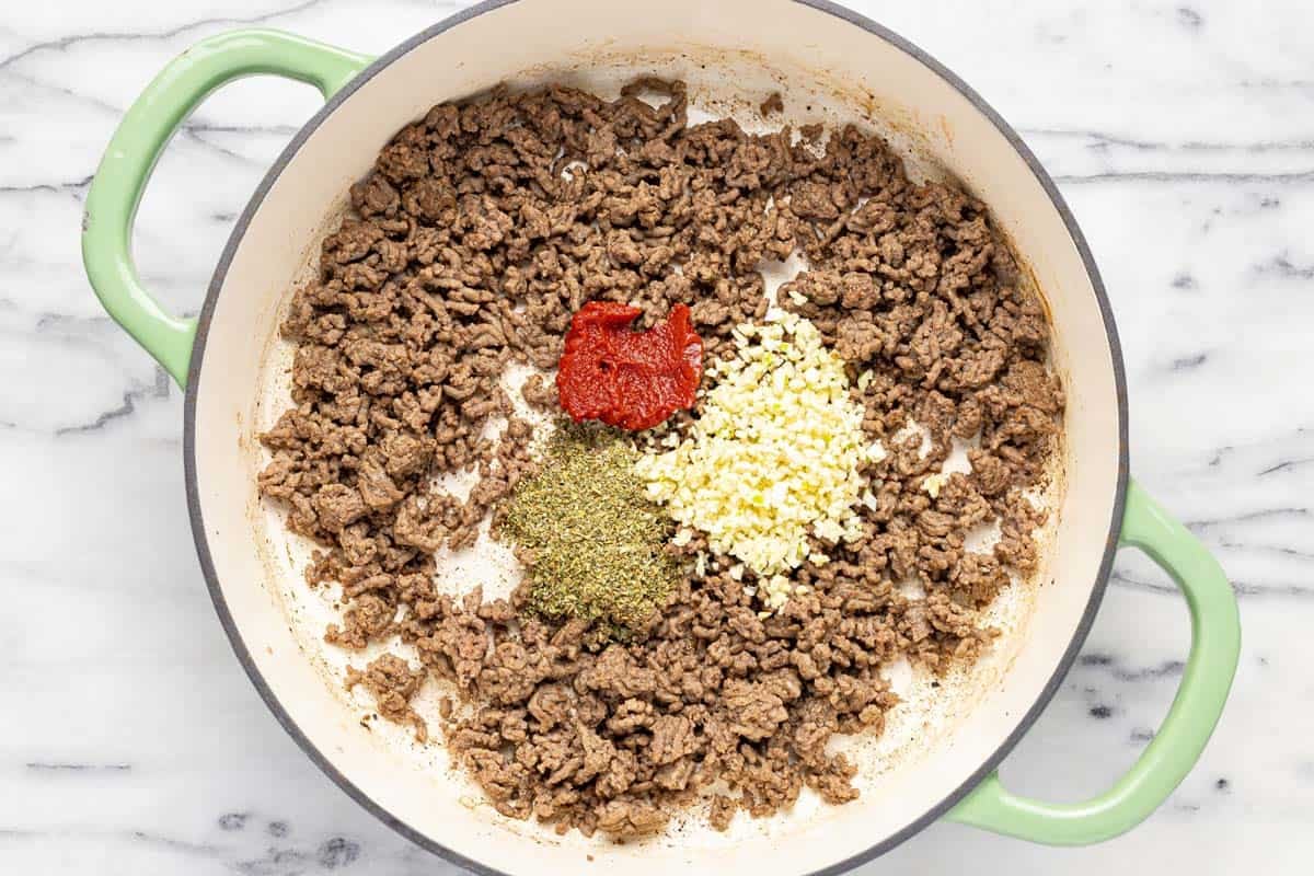 Cooked ground beef with tomato paste, herbs, and minced garlic in a large pan. 