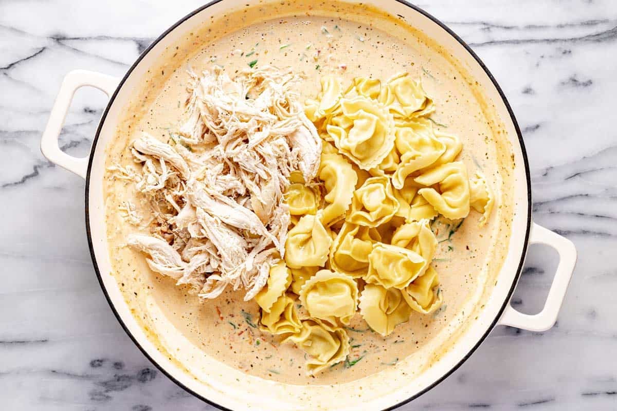 Shredded chicken and tortellini added to a large pan of creamy sauce. 
