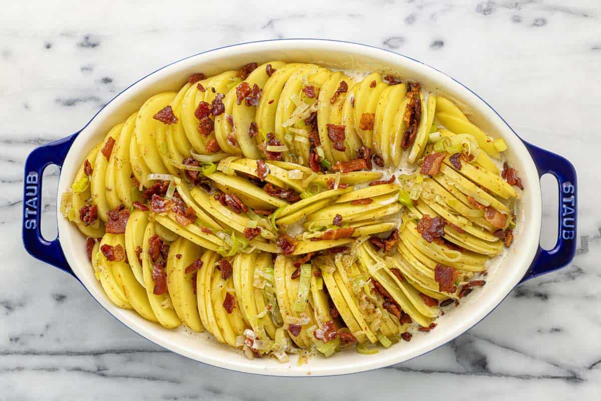 Sliced potatoes in a casserole dish with sauce, bacon, and leeks. 