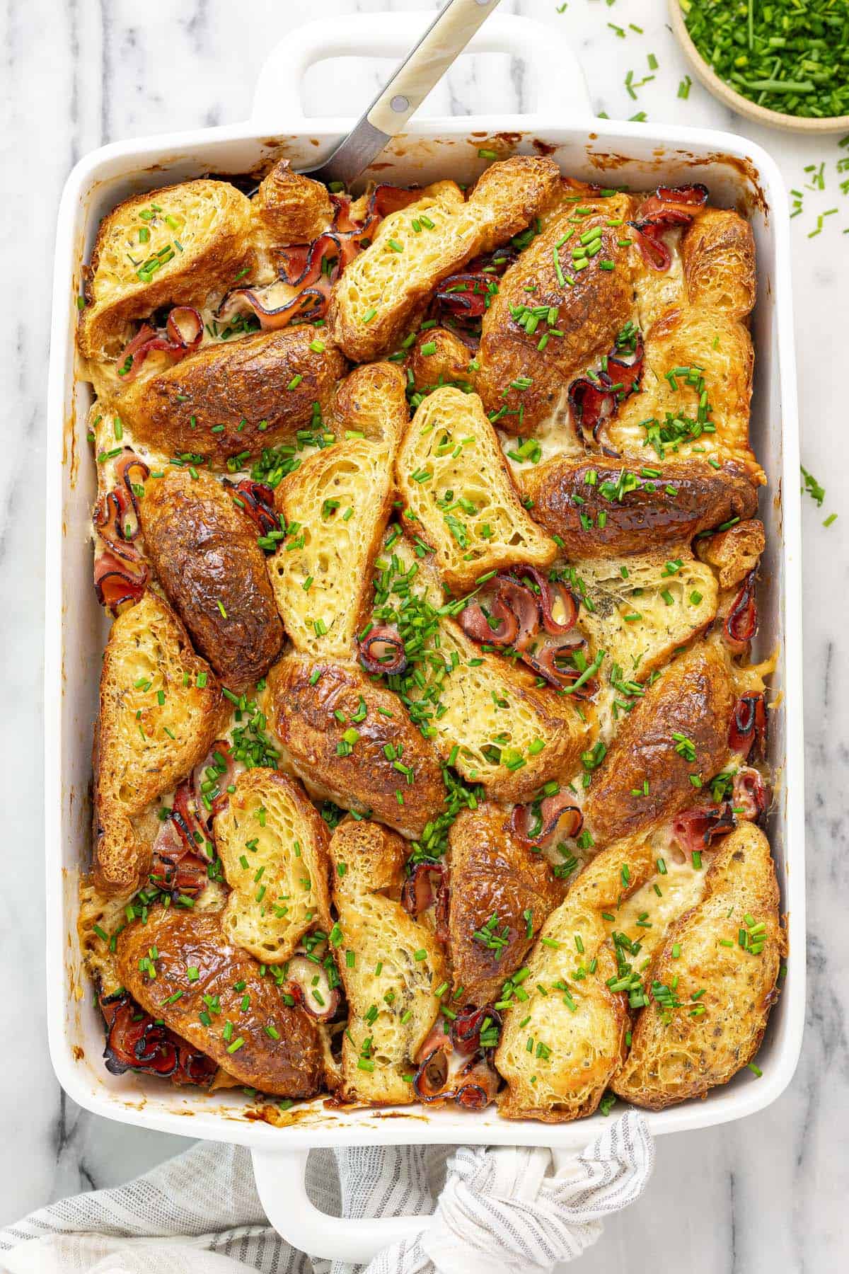 Baking dish filled with croissants, ham, and cheese in an egg custard bake garnished with chives.