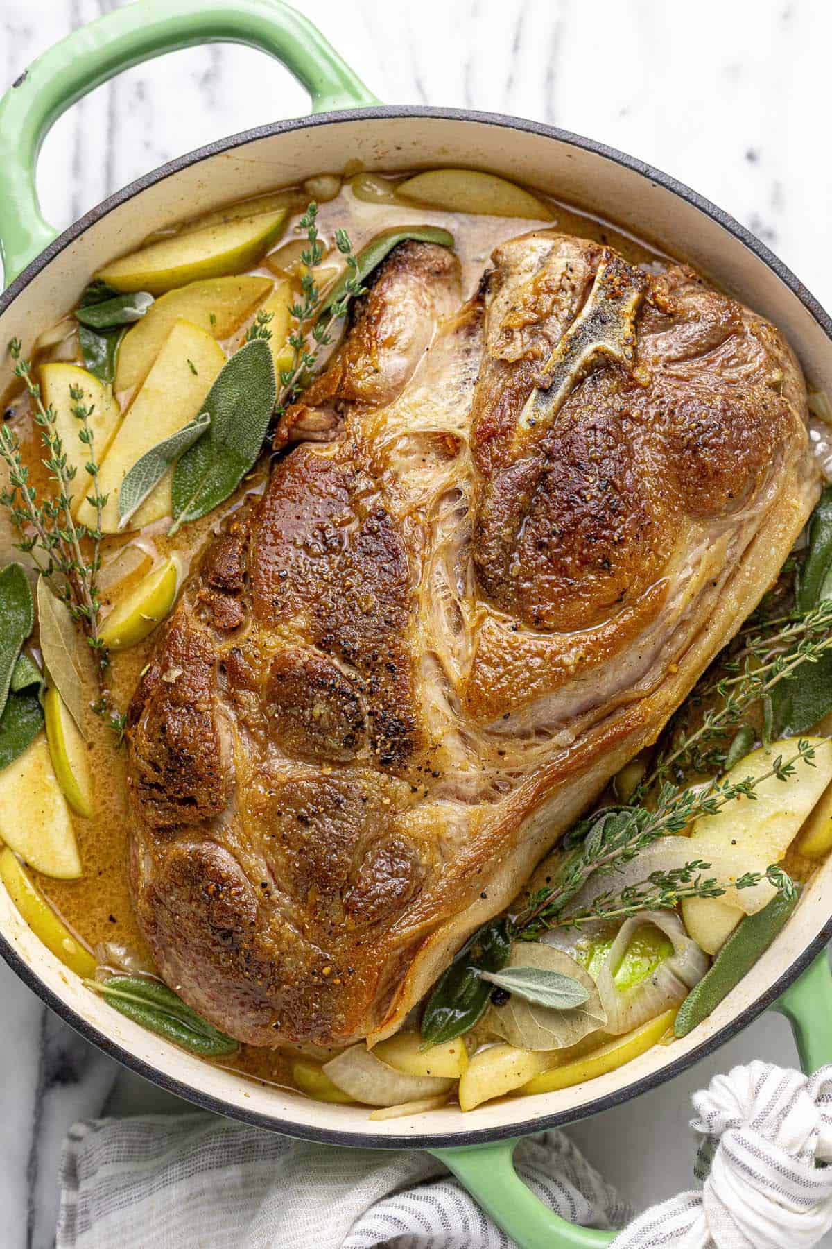 Seared pork shoulder in a pan with herbs, apples, and onions in broth. 