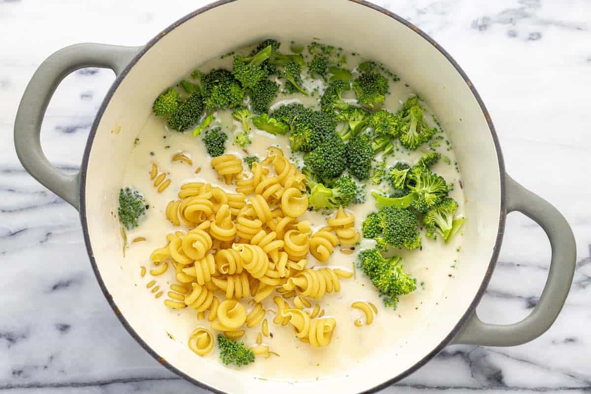 Broccoli and uncooked pasta added to a large pot of soup.