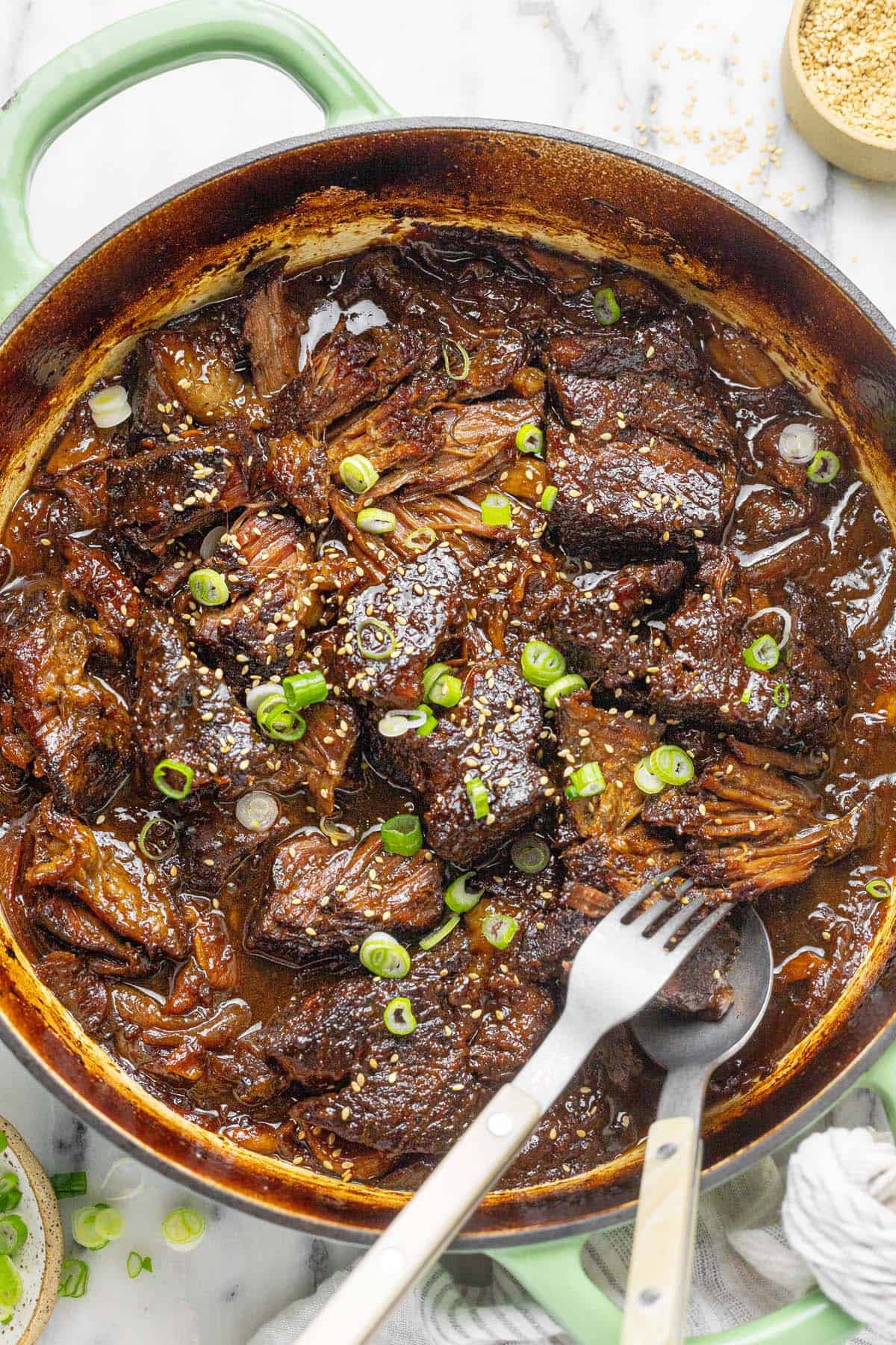 Shredded braised beef chuck roast in a large pan with sesame seeds and sliced green onion.