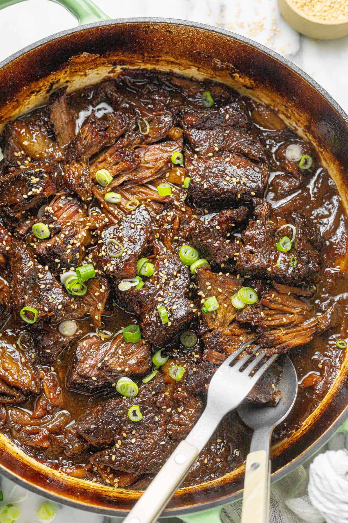 Shredded braised beef chuck roast in a large pan with sesame seeds and sliced green onions.