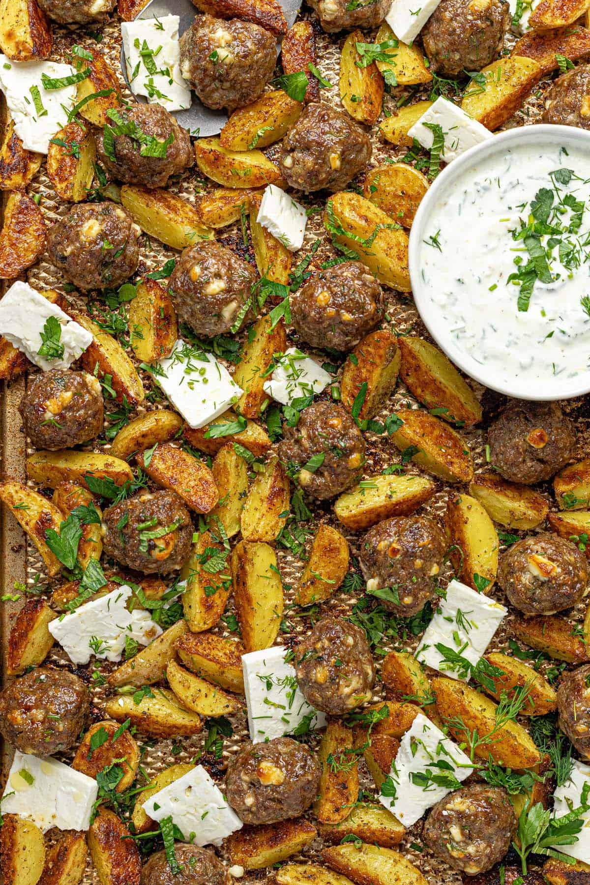 Close up of a sheet pan filled with roasted potatoe, Greek meatballs, sliced feta, and yogurt sauce.