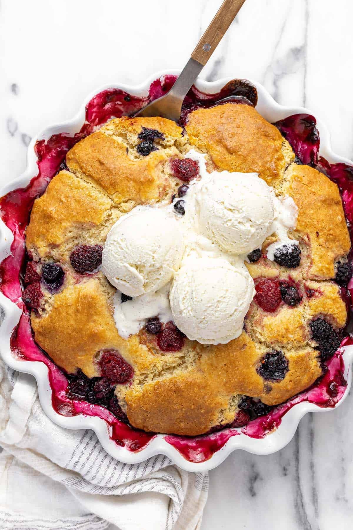Large pie dish filled with homemade triple berry cobbler topped with ice cream.