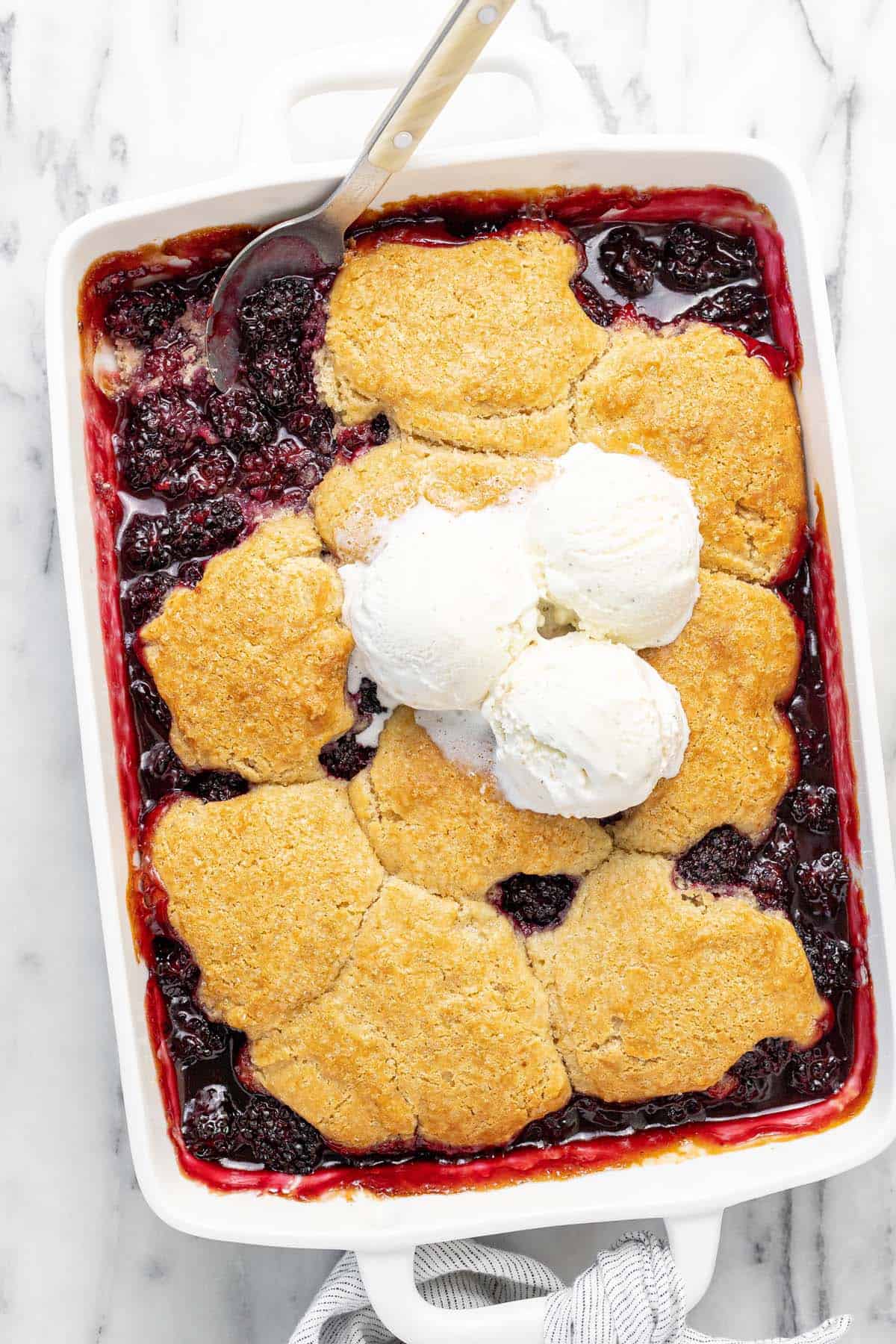 Blackberry cobbler in a large baking dish topped with vanilla ice cream.