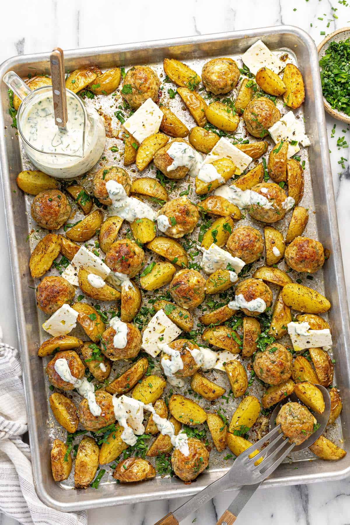 Sheet pan filled with Greek chicken meatballs and potatoes with sliced feta cheese.