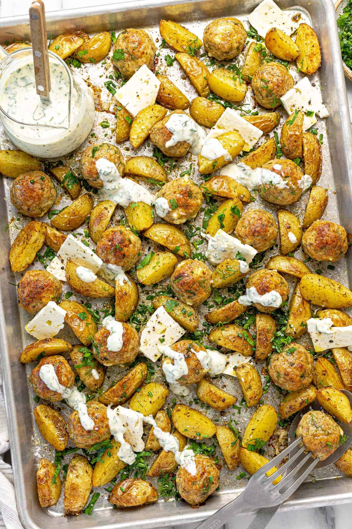 Sheet pan filled with Greek chicken meatballs and potatoes with sliced feta cheese.