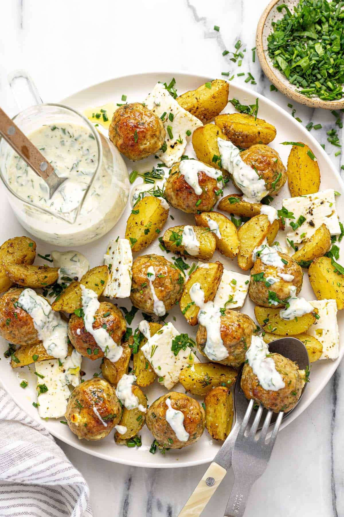 Large plate filled with Greek chicken meatballs and roasted potato wedges drizzled with yogurt sauce.