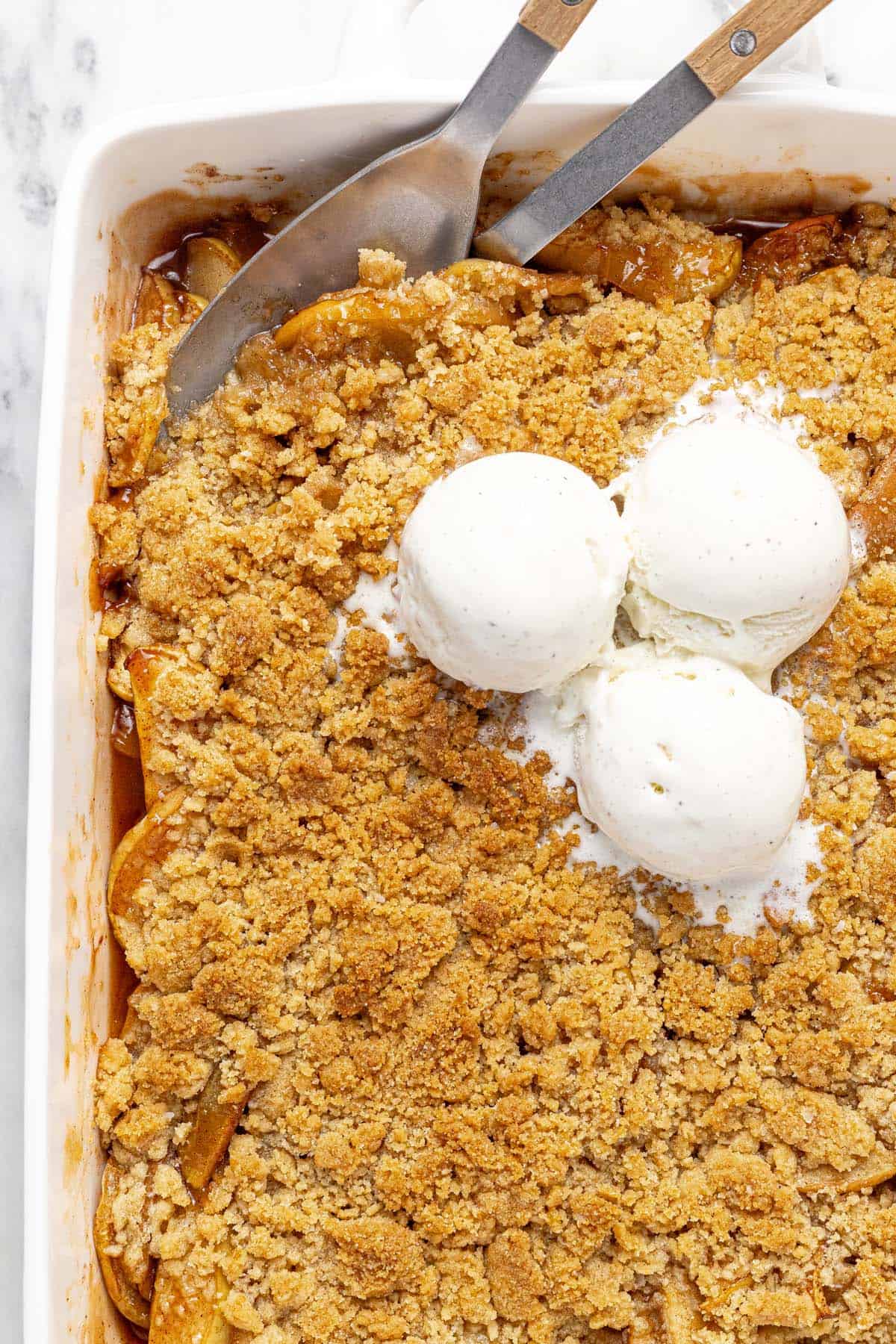 Close up of a pan filled with homemade apple crumble topped with scoops of vanilla ice cream.
