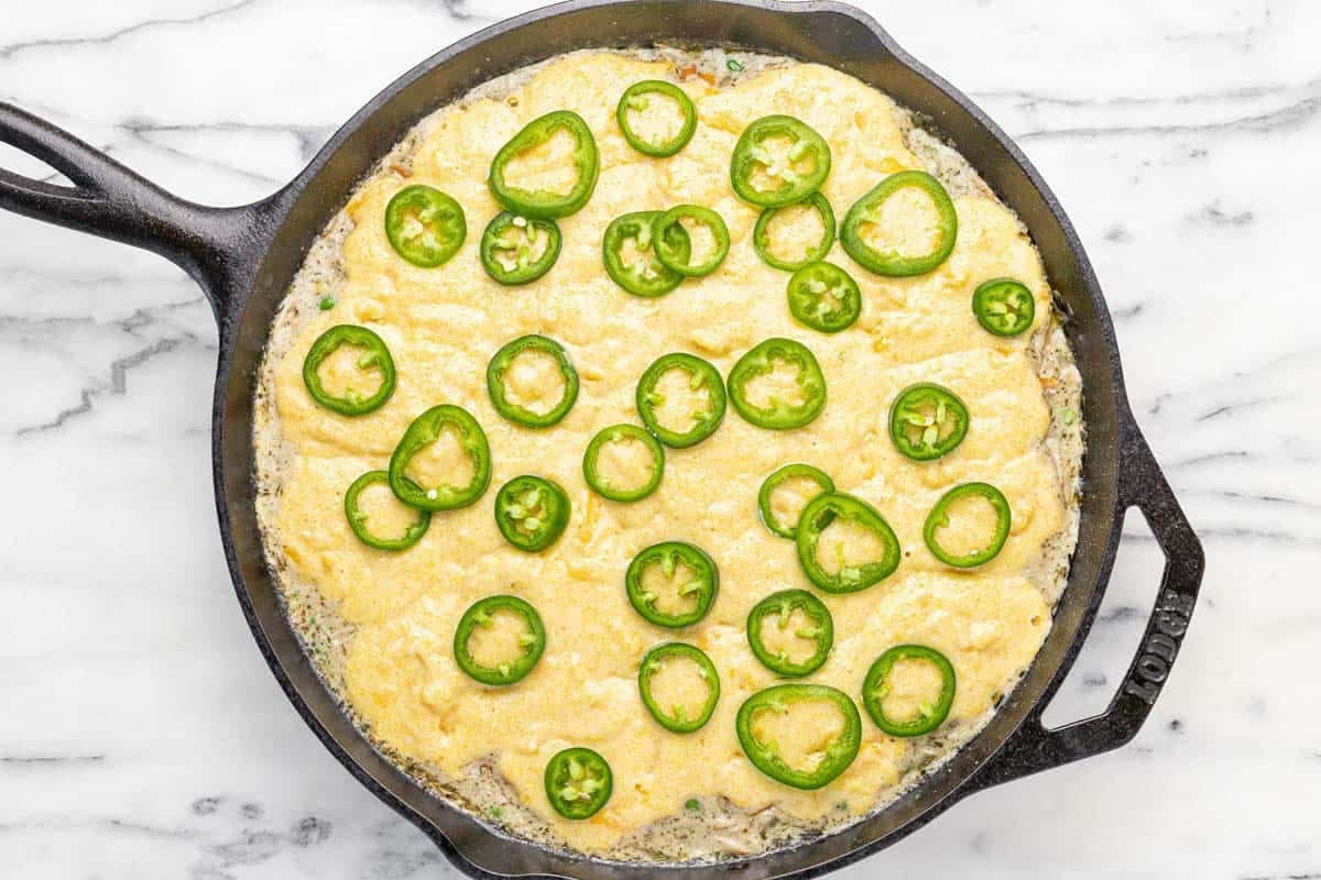 Cornbread chicken pot pie topped with jalapenos in a large cast iron skillet ready for the oven.